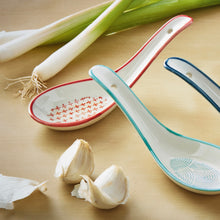 Load image into Gallery viewer, White stoneware geo soup spoons with red, teal, and blue rim accents and geometric patterned interiors, displayed on a neutral background with green onions and garlic cloves.   