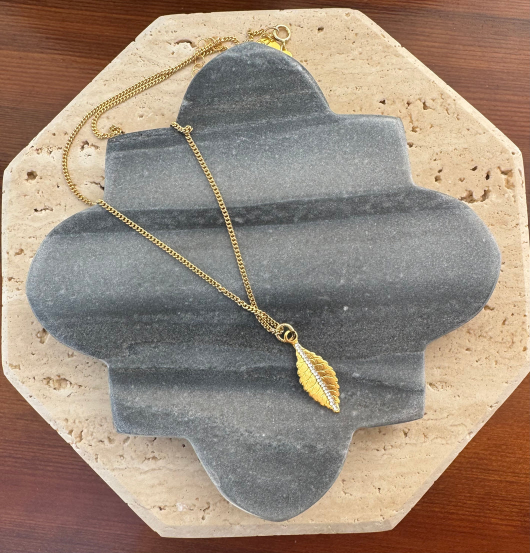 Gold leaf pendant necklace featuring a delicate gold chain and a central row of sparkling crystals, displayed on a quatrefoil-shaped gray stone tile layered over a beige travertine slab on a wooden surface.  