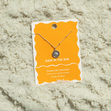 Load image into Gallery viewer, Sterling silver-dipped necklace featuring a small sun charm pendant and a chain with delicate beaded accents, displayed on a golden yellow &quot;Bask in the Sun&quot; Ocean Life Collection card by Lucky Feather with a white wavy border, shown resting on white textured sand. 