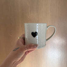 Load image into Gallery viewer, Hand holding a white stoneware mug with a black heart design against a wood background. 