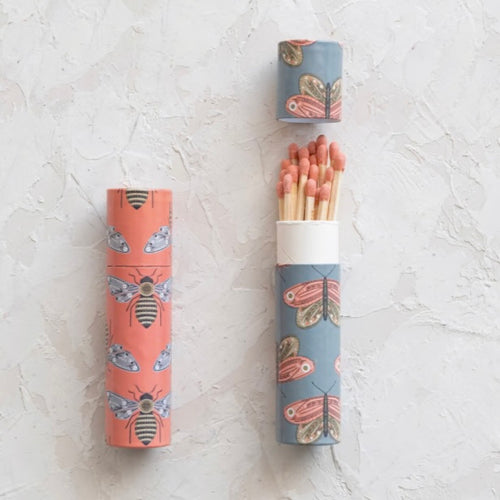 Two tube boxes of matches with an insect pattern. One tube is pink with a bee design. The second tube is blue with a butterfly pattern and is open revealing the long red tipped matches. Both are against a white textured  background. 