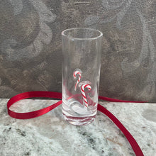 Load image into Gallery viewer, ZODAX Holiday Shot Glasses, lifestyle image of a tall clear shot glass with a thick base and raised red-and-white striped candy cane motifs, resting on a gray marble surface with a red ribbon and a patterned gray background.
