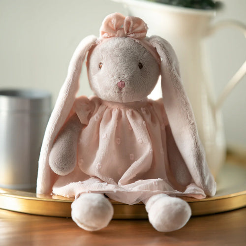 Plush white bunny doll with long ears wearing a soft peach-colored dress with subtle embroidered dots and a matching ruffled headband, seated on a gold rimmed round tray with a blurred background.