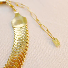 Load image into Gallery viewer, Close-up of the gold-plated brass herringbone chain necklace showing the polished links, end caps, and adjustable extender chain with a small "West Native" tag, displayed flat on an off-white textured surface.  