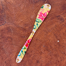 Load image into Gallery viewer, A yellow canapé knife with a pink, green, and white floral design . On a brown wooden surface. 