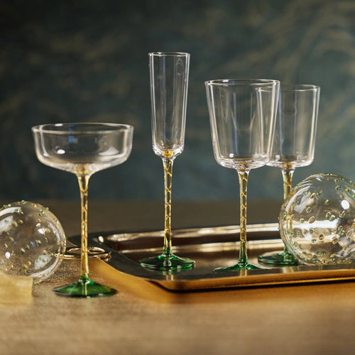 Assortment of clear glassware featuring three styles with amber stems and green bases displayed on a gold tray. The glassware includes a champagne flute, two red wine glasses, and a white wine glass, shown with decorative glass spheres on a golden surface with a dark background. 