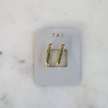 Load image into Gallery viewer, Gold-plated baguette earrings with slim rectangular emerald green stones on a light gray TAI jewelry card, displayed on a white marble background.   