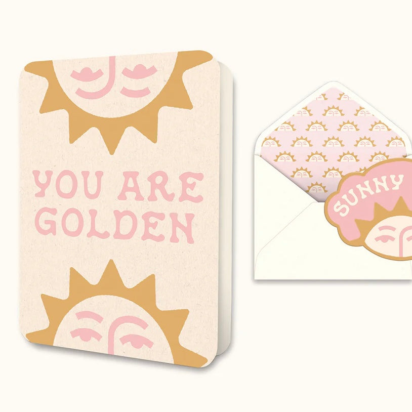 Cream-colored greeting card with stylized golden sun faces and pink text reading 