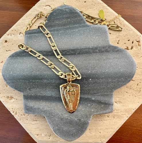 Gold paper clip chain necklace with a raised Archangel pendant, displayed on a quatrefoil-shaped gray stone tile layered over a beige travertine slab on a wooden surface.  
