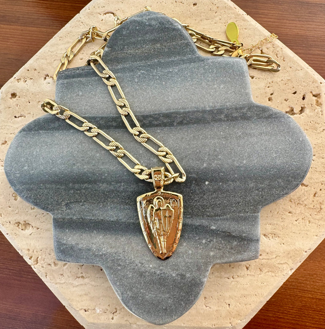 Gold paper clip chain necklace with a raised Archangel pendant, displayed on a quatrefoil-shaped gray stone tile layered over a beige travertine slab on a wooden surface.  