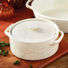 Load image into Gallery viewer, Harvest Round Brie Baking Dish, round warm off-white stoneware dish with two smooth open handles, matching embossed leaf designs around the exterior and fitted lid, and a smooth low round lid knob, displayed on a wooden surface with matching baking dish in the background. 