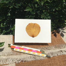 Load image into Gallery viewer, Greeting card with a brown pressed flower petal and various colored ink pens on a beige and white blanket, with bushes in the background.