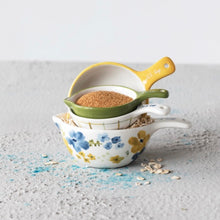 Load image into Gallery viewer, A set of nesting  ceramic measuring cups. Largest and first cup features a floral design with oats inside, 2nd  is white with brown stripes, 3rd cup  is the smallest and an olive green color with brown powder, and a yellow measuring cup standing behind the three other cups. Cups on a white textured surface , dusted with oats. Against a white background.