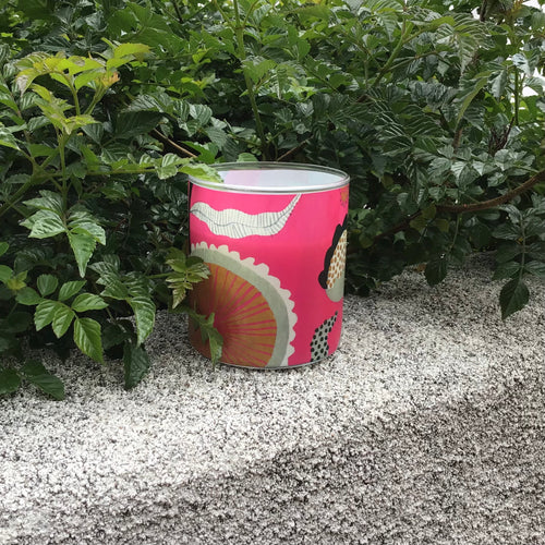 Glass candle with pink and whimsical designs on a concrete surface and bushes in the background. 