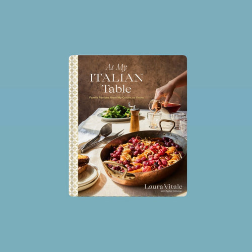 Book cover of At My Italian Table by Laura Vitale featuring a rustic dinning scene with a large copper pan of roasted vegetables and bread on a linen-covered table, surrounded by plates, utensils, a carafe of wine, and a green salad in the background. Book against a blue background. 