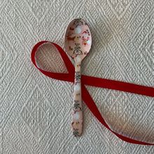 Load image into Gallery viewer, Enameled Holiday Spoons | Four Styles, lifestyle photo of a stainless steel spoon with a pale pink enamel design featuring Santa Claus, a snowman, a reindeer, gifts, and holiday decorations, displayed with a red ribbon on a cream textured fabric surface. 