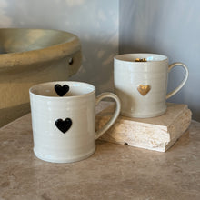 Load image into Gallery viewer, Two white stoneware mugs with heart designs, one with a black heart and one with a gold heart, displayed on a tabletop.   