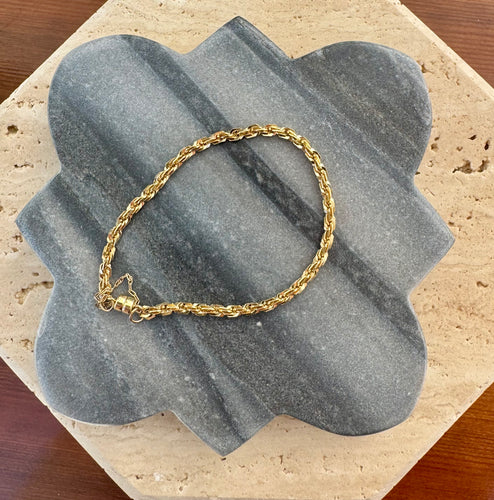 Gold rope chain bracelet displayed on a gray quatrefoil-shaped stone tile layered over a beige travertine slab on a wooden surface.