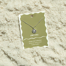 Load image into Gallery viewer, Sterling silver-dipped necklace featuring a small sea turtle charm pendant and a chain with delicate beaded accents, displayed on an olive green &quot;Stay Chill&quot; Ocean Life Collection card by Lucky Feather with a white wavy border, shown resting on white textured sand. 