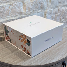 Load image into Gallery viewer, Closed Byrdeen gift box with a white lid and floral patterned sides, displayed on a round wooden table against a textured gray stone wall. 