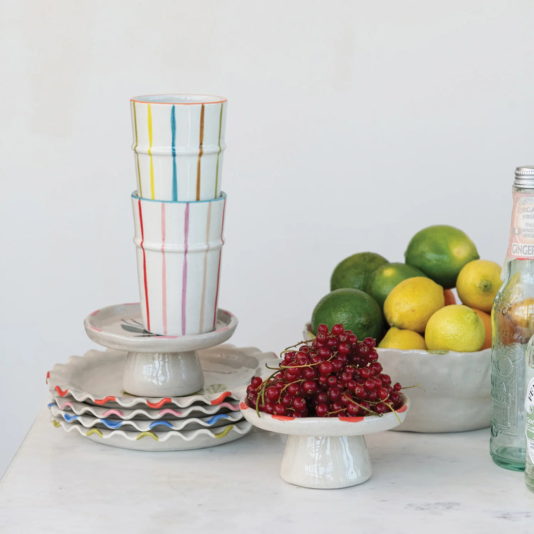 A set of multicolored striped cups sitting on a stack of white dishes with colored accents. Alongside, a dish of fruit, a bowl of lemons and limes, and clear glass bottles. On a white surface with a white background.