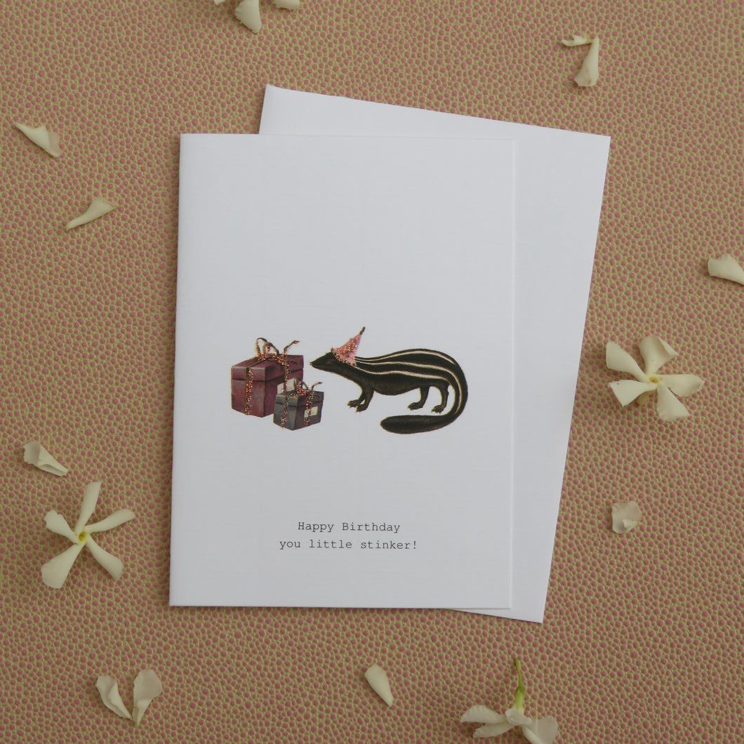 Greeting card with skunk and gifts illustration and birthday message on a textured surface with white flowers. 