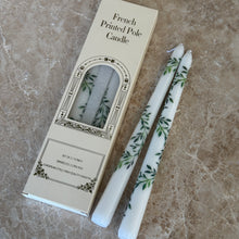 Load image into Gallery viewer, Two white taper candles with green botanical designs on a brown marble surface, next to their packaging. 