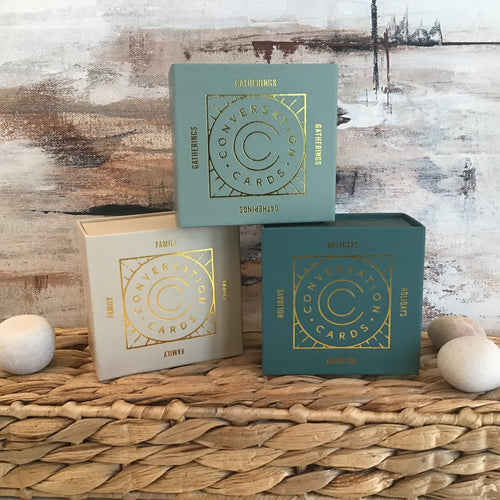 Three boxes stacked in a pyramid on a braided wicker stand with stones to either side. Boxes are cream, light blue, or teal with a gold foil circular design that reads 