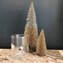 Load image into Gallery viewer, A glass tumbler with a snowman design. The snowman is white with a black top hat, a red scarf, an orange dot for the carrot nose, and black dots for the eyes and buttons. Tumbler and two frosted decorative Christmas trees on a cream and brown marble slab. Against a blue background. 