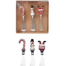 Load image into Gallery viewer, Set of three holiday cheese spreaders with glass handles featuring a candy cane, a snowman, and Santa&#39;s legs in a chimney, displayed in a brown packaging box. Matching holiday spreaders below box. All against a white background.   