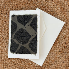 Load image into Gallery viewer, Colby Cards, lifestyle image of a white Lokta paper greeting card featuring detailed black leaf-vein prints on a gray panel with softly textured, uneven edges, displayed with a white envelope on a brown woven mat.