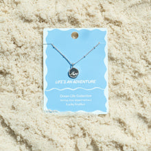 Load image into Gallery viewer, Sterling silver-dipped necklace featuring a small wave charm pendant and a chain with delicate beaded accents, displayed on a blue &quot;Life&#39;s an Adventure&quot; Ocean Life Collection card by Lucky Feather with a white wavy border, shown resting on white textured sand. 