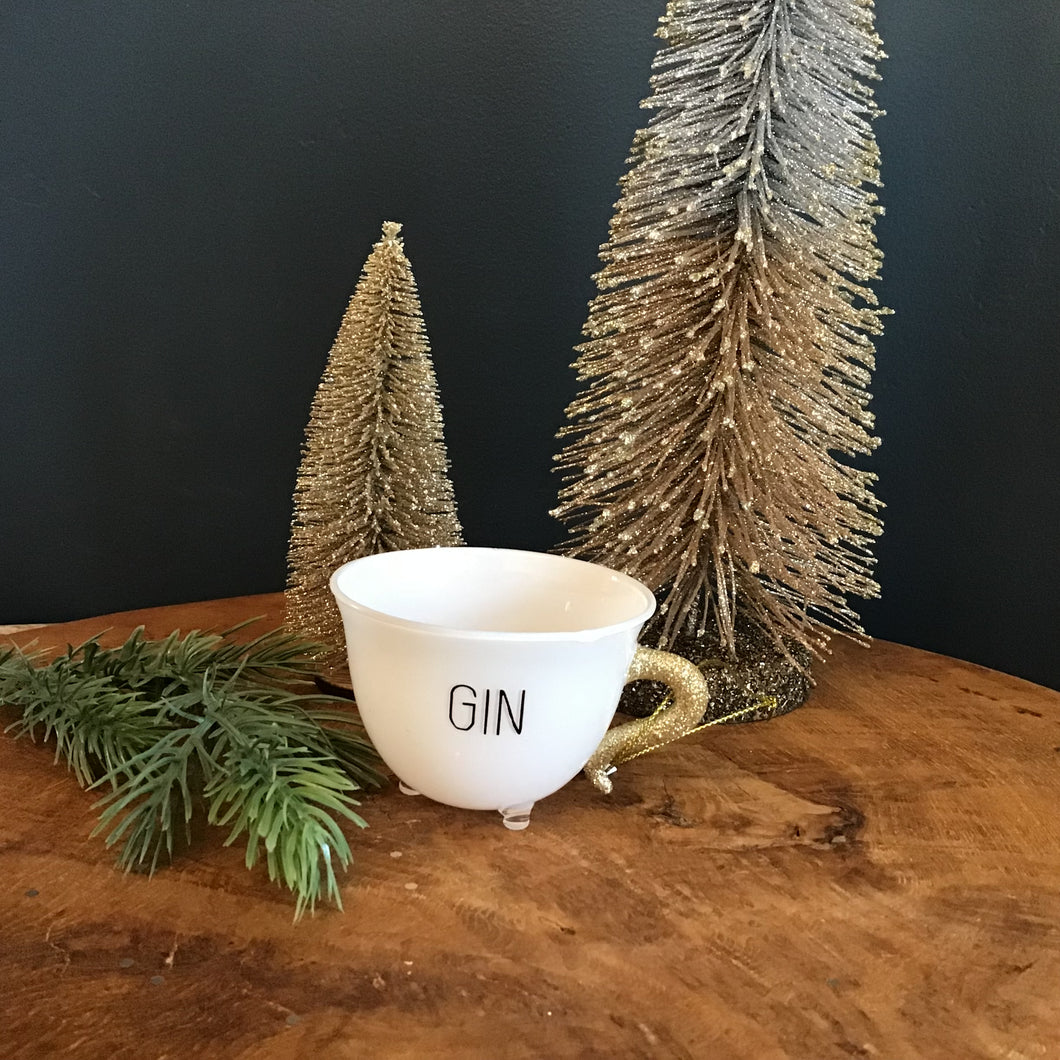 A white teacup ornament with a gold glitter handle, a clear footed bottom,  and 
