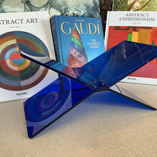 Load image into Gallery viewer, Blue Acrylic Book Holder, transparent blue acrylic book holder with interlocking X-shaped design, displayed on beige textured surface with books in the background. 