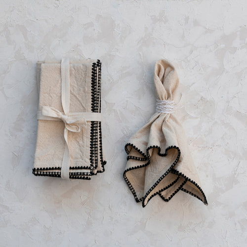 Beige square cotton napkins with black trim. A set folded neatly and tied in a white ribbon , and a single napkin with a white string napkin holder, displayed on a white textured surface.