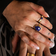 Load image into Gallery viewer, Model&#39;s manicured hands placed together wearing a double-gold-plated brass ring with an oval lapis stone and small CZ stone accents, along with coordinating rings, shown against a softly lit background. 