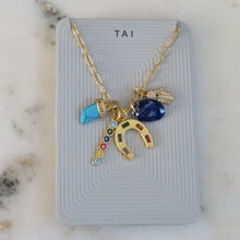 Load image into Gallery viewer, Gold-tone charm necklace displayed on a light gray TAI jewelry card featuring a turquoise-colored horn charm, a faceted deep blue stone charm, a gold horseshoe charm with multi-colored inlays, a small vertical multi-colored evil eye charm, and a pave hamsa hand charm.  