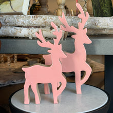 Load image into Gallery viewer, Pink Dancing Deer, lifestyle image of two pink mango wood deer in small and large sizes, each with branching antlers, one raised front leg, and natural wood edges, standing on a round light-colored surface with a black rim in front of stacked birch logs.