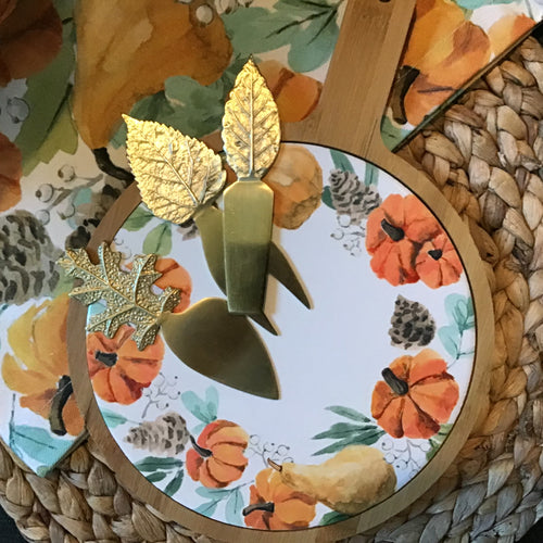 White enamel plate with orange pumpkin , gourd, brown pinecone, and green leaf design on a round, wooden, brown tray with a handle. Three gold cheese utensils with a leaf handle on the enamel plate. Tray next to a matching table runner , on a woven mat.