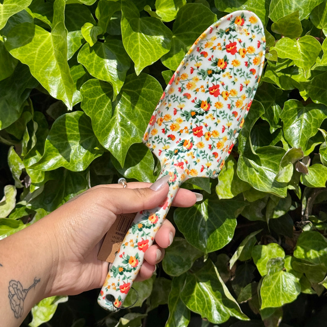 Hand holding a white floral-patterned garden shovel against a green leafy background
