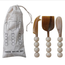 Load image into Gallery viewer, Horn Cheese Knives Set, three tortoise-toned natural horn cheese knives, white resin bead handles, and natural-colored drawstring storage pouch with illustrated black cheese knife outline design against white background. 