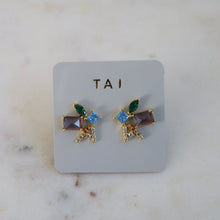 Load image into Gallery viewer, Pair of gold-plated mixed-shaped cluster stud earrings with multicolored crystals, displayed on a light gray TAI jewelry card against a white marble background. 