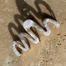 Load image into Gallery viewer, Pearl white wavy claw clip with gold metal hardware displayed on a beige travertine surface with soft shadows.