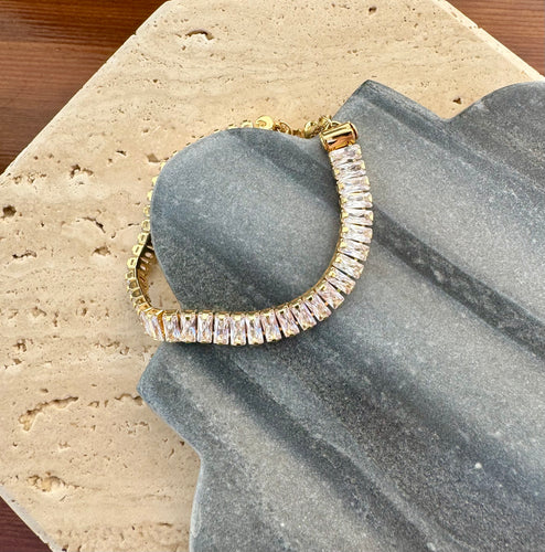 Bracelet featuring a row of rectangular clear stones in a shiny warm toned metal, displayed in a curved position on a quatrefoil-shaped gray stone tile layered over a beige travertine slab on a wooden surface. 