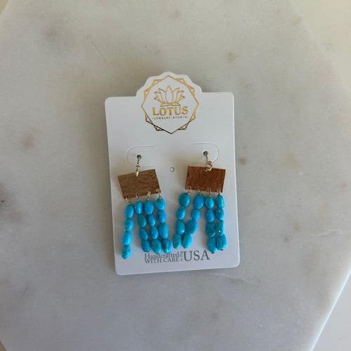Pair of earrings each with a thing gold sheet and four strands of blue beads attached. Earrings fixed to a white piece of cardboard flat laid on a white marble background.