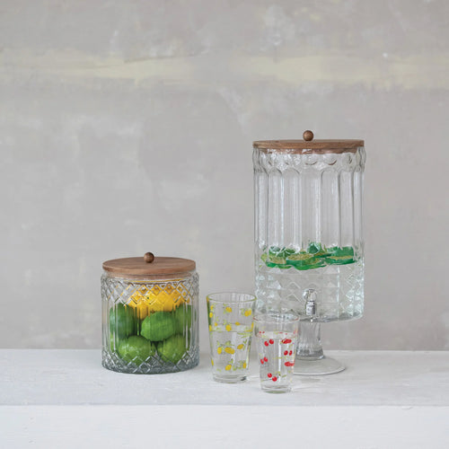 A set of two glass canisters with wood lids, one smaller than the other. The smaller canister contains green limes and yellow lemons, and the larger one has green candies. Alongside of yellow and red shot glasses. On a white surface, with gray wall behind. 