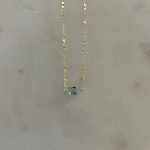 Gold chain necklace with a square aquamarine gem at the center. White marble background. 