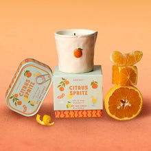 Load image into Gallery viewer, Soy wax candle in a white ceramic cup container with small orange motif, displayed on the Citrus Spritz product box with matching illustrated tin candle and fresh orange slices, against a peach background.