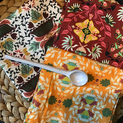 Three cotton kitchen towels in burgundy, orange, and cream, each with floral patterns , displayed on a brown woven surface with a white long handle spoon resting on top.  