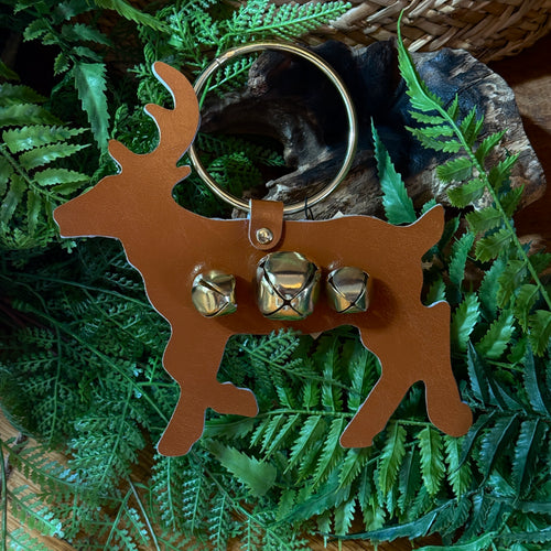 Brown metal deer shape ornament with three gold jingle bells. A silver metal ring attached to the deer for hanging. Lying on a bed of green ferns and a charred log . Brown woven basket partially visible in top right corner. 